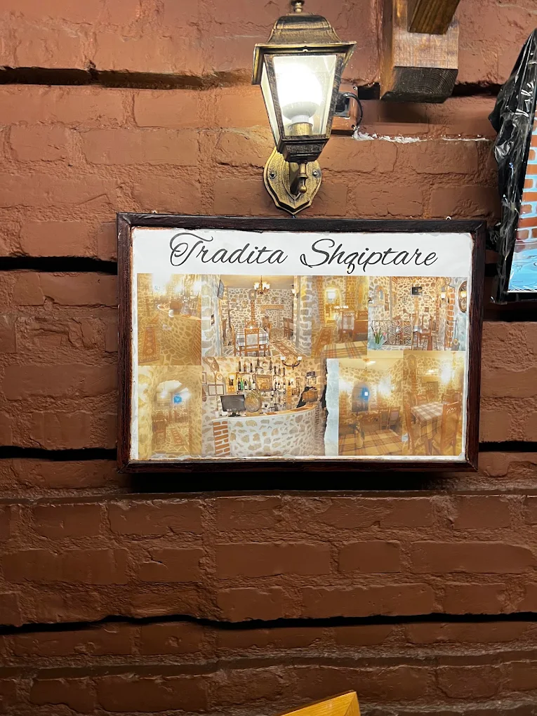 Tradita Shqiptare nameplate and restaurant photographs on the brick wall beneath a lantern.